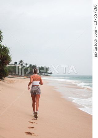 Beach Run Alone. Morning Jog Along Sandy Shore. Lone Woman Exercises On Beach With Footprints In Sand 137557260
