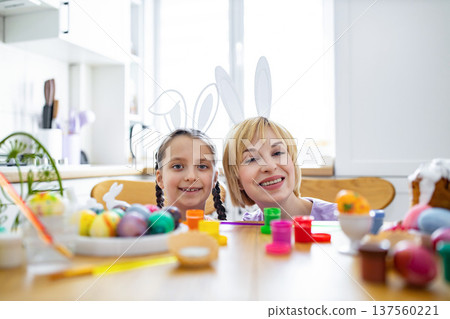 A mother and daughter wearing bunny ears peek over a table filled with colorful Easter eggs and painting supplies, ready for holiday activities A mother and daughter wearing bunny ears peek over a table filled with colorful Easter eggs and painting supplies, ready for holiday activities 137560221