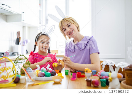 A mother and daughter happily paint colorful Easter eggs together at a kitchen table, wearing bunny ears and surrounded by festive decorations A mother and daughter happily paint colorful Easter eggs together at a kitchen table, wearing bunny ears and surrounded by festive decorations 137560222