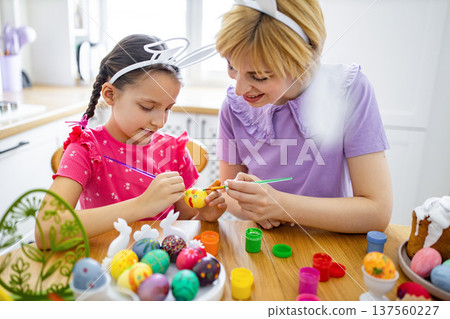 A mother and her daughter wearing bunny ears are happily painting colorful Easter eggs together at a kitchen table, surrounded by art supplies and decorated eggs A mother and her daughter wearing bunny ears are happily painting colorful Easter eggs together at a kitchen table, surrounded by art supplies and decorated eggs 137560227