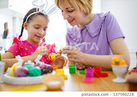 A mother and her daughter are happily painting colorful Easter eggs together at a wooden table, creating festive decorations for the holiday A mother and her daughter are happily painting colorful Easter eggs together at a wooden table, creating festive decorations for the holiday 137560228