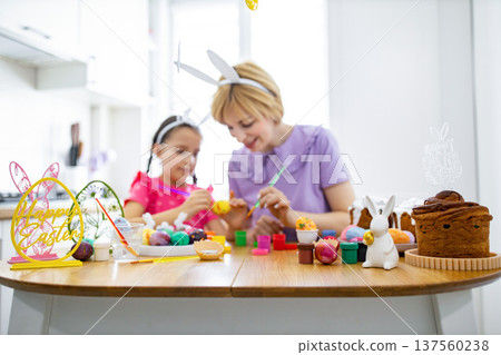 A mother and daughter happily paint colorful Easter eggs together at a wooden table, surrounded by festive decorations and traditional cakes A mother and daughter happily paint colorful Easter eggs together at a wooden table, surrounded by festive decorations and traditional cakes 137560238