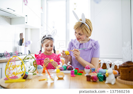 A mother and her daughter wearing bunny ears happily paint colorful Easter eggs together at a kitchen table, surrounded by festive decorations and cakes 137560256