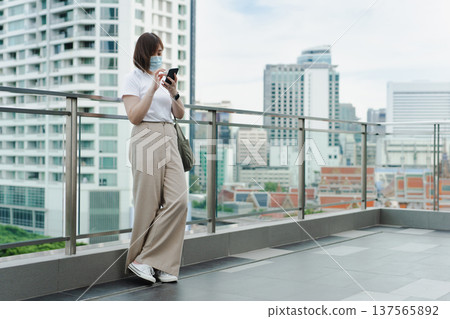 solo asian woman wear protective mask during use phone and relax at rooftop with city background solo asian woman wear protective mask during use phone and relax at rooftop with city background 137565892