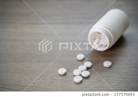 White round pills spilling from a plastic bottle on wooden table, close up healthcare background. Concept of medication, prescription drugs, pharmacy, medical treatment and dosage control. White round pills spilling from a plastic bottle on wooden table, close up healthcare background. Concept of medication, prescription drugs, pharmacy, medical treatment and dosage control. 137570893
