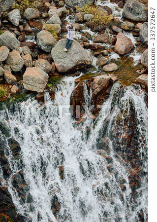 A photographer girl sits on a rock at the edge of the Battery waterfall in Teriberka, Arctic landscape with a view of a flowing waterfall in the northern fjord, Arctic, Russia, Murmansk region 137572647