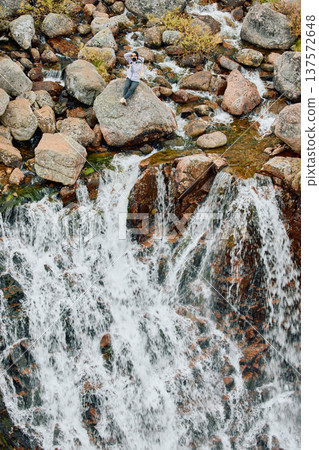 A photographer girl sits on a rock at the edge of the Battery waterfall in Teriberka, Arctic landscape with a view of a flowing waterfall in the northern fjord, Arctic, Russia, Murmansk region A photographer girl sits on a rock at the edge of the Battery waterfall in Teriberka, Arctic landscape with a view of a flowing waterfall in the northern fjord, Arctic, Russia, Murmansk region 137572648