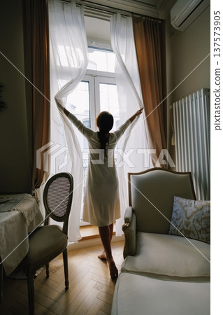Woman opening curtains in morning light Woman opening curtains in morning light 137573905