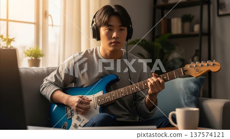 Young Asian man practicing electric guitar with headphones in modern living room during daytime music lesson 137574121