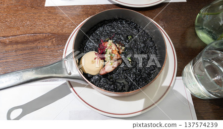 Mediterranean squid ink rice served in a metal pan with tender squid and creamy sauce, styled as a rich black seafood dish with elegant restaurant presentation. Mediterranean squid ink rice served in a metal pan with tender squid and creamy sauce, styled as a rich black seafood dish with elegant restaurant presentation. 137574239
