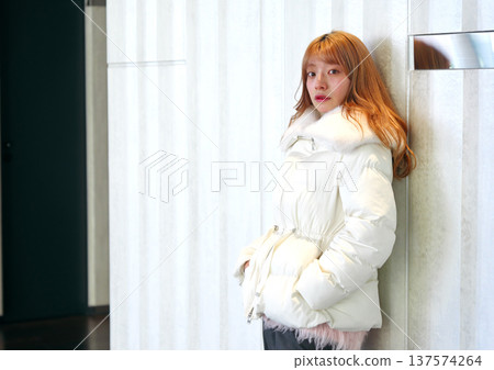 A female model leaning against a white wall. A sophisticated winter indoor portrait with a clean white outerwear. 137574264