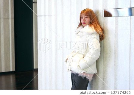 一位女模特兒倚靠在白牆上。這是一張精緻的冬季室內肖像照，她穿著乾淨的白色外套。 137574265