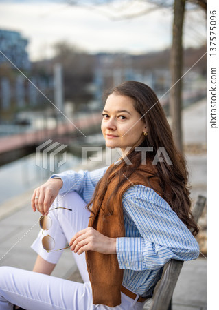 Elegant young woman in old money style sitting on bench near pier in Prague holding sunglasses in early spring. Understated luxury and timeless European femininity concept. 137575096