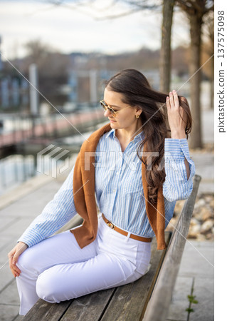 Elegant young woman in old money style sitting on bench near pier in Prague wearing sunglasses in early spring. Quiet luxury lifestyle and refined European elegance concept. 137575098