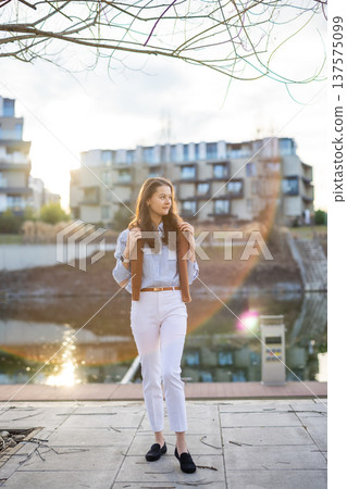 Elegant young woman in old money style standing by riverside near Prague pier in early spring sunlight. Quiet luxury aesthetic and confident European lifestyle concept. 137575099