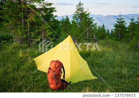 Yellow tourist tent on grassy hillside, accompanied by orange backpack and camera tripod. Stunning view of distant blue mountains under clear sky, perfect for peaceful camping adventure. 137576673