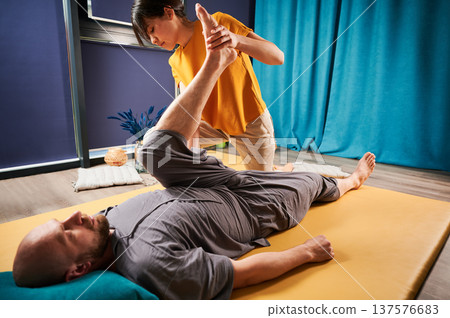 Masseuse making thai yoga massage. Woman therapist doing traditional massage treatment, stretching male patient's body. Bald client lying on mat, enjoying hand massage in calm atmosphere. Masseuse making thai yoga massage. Woman therapist doing traditional massage treatment, stretching male patient's body. Bald client lying on mat, enjoying hand massage in calm atmosphere. 137576683