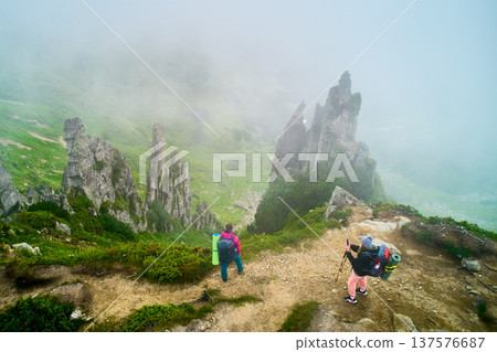 Aerial view of two hikers stand at edge of misty mountain ridge, surrounded by towering rock formations and lush green vegetation. One hiker taking photo while other looks out over foggy landscape. Aerial view of two hikers stand at edge of misty mountain ridge, surrounded by towering rock formations and lush green vegetation. One hiker taking photo while other looks out over foggy landscape. 137576687