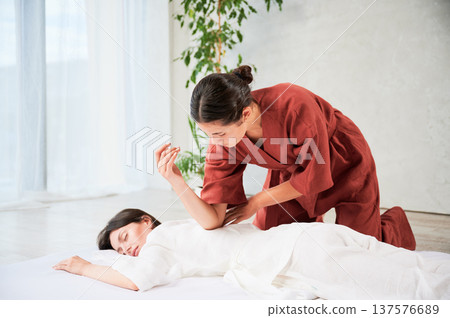 Masseuse making thai yoga massage. Woman therapist doing traditional massage treatment for female patient using elbow. Client lying on mat, meditating, enjoying hand massage in calm atmosphere. 137576689