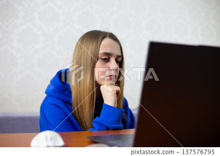 Young woman in blue hoodie sits at desk chin on fist intently watching Football World Cup. National team match live stream on laptop screen focused expression indoor home setting Young woman in blue hoodie sits at desk chin on fist intently watching Football World Cup. National team match live stream on laptop screen focused expression indoor home setting 137576795