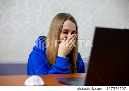 Blonde girl laughs at funny football moment on laptop, covering mouth with hand. Amusing sports blooper or unexpected play during match makes her giggle. Entertainment at home 137576797