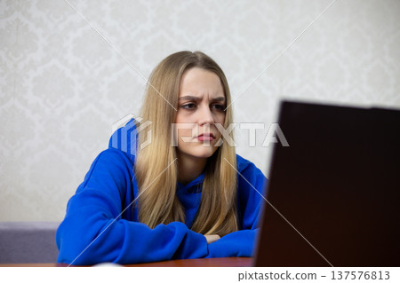 Young woman with straight blonde hair in a blue hoodie leans forward at a desk, focusing on a laptop screen in a bright room featuring light-colored patterned wallpaper Young woman with straight blonde hair in a blue hoodie leans forward at a desk, focusing on a laptop screen in a bright room featuring light-colored patterned wallpaper 137576813