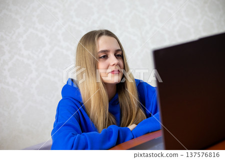 Young blonde woman smiles gently while listening to motivational video on laptop. Inspirational speaker uplifts her mood, encourages personal growth. Positive online content at home 137576816