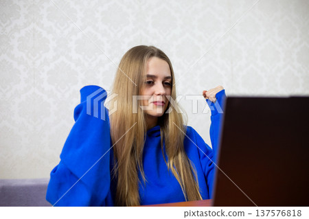 Excited blonde girl celebrates national football team victory at World Cup. She raises clenched fists in triumph while watching winning match on laptop. Joyful fan at home 137576818