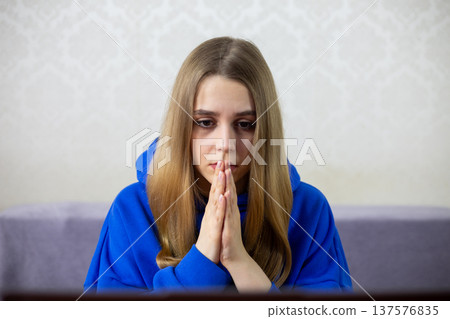 Young woman in blue hoodie prays for loan approval while checking bank account online. Hands clasped in hope for mortgage or credit acceptance. Financial concept Young woman in blue hoodie prays for loan approval while checking bank account online. Hands clasped in hope for mortgage or credit acceptance. Financial concept 137576835