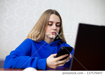 Young blonde woman uses laptop and smartphone simultaneously at home. Surrounded by digital devices, she represents modern generation immersed in technology and constant connectivity Young blonde woman uses laptop and smartphone simultaneously at home. Surrounded by digital devices, she represents modern generation immersed in technology and constant connectivity 137576847