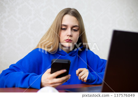 Young woman checks smartphone for verification code while shopping online on laptop. She confirms payment or login securely at home. Two factor authentication and digital security concept 137576849