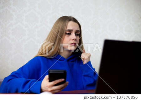 Young woman with long blonde hair wearing a blue hoodie holds a smartphone while gazing at a laptop screen, seated at a wooden desk with a light-colored patterned wall behind her Young woman with long blonde hair wearing a blue hoodie holds a smartphone while gazing at a laptop screen, seated at a wooden desk with a light-colored patterned wall behind her 137576856