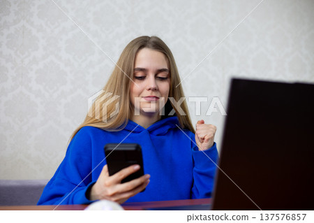 Happy blonde girl celebrates winning sports bet on smartphone. She clenches fist in victory while checking successful wager on betting app. Joyful gambler at home 137576857