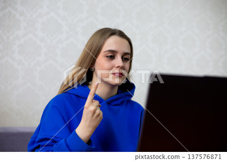 Excited blonde football fan celebrates first goal with index finger pointed at laptop screen. She cheers for her team during live match broadcast. Joyful reaction at home 137576871