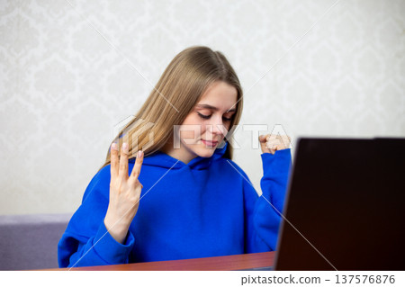Blonde football fan celebrates third goal with three fingers raised toward laptop. She witnesses hat trick or decisive score during live match. Intense excitement at home 137576876