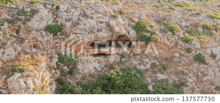 Ancient rocky cave entrance in stone cliff, geological formation with dry vegetation under bright daylight, archaeological site and wild nature landscape Ancient rocky cave entrance in stone cliff, geological formation with dry vegetation under bright daylight, archaeological site and wild nature landscape 137577730