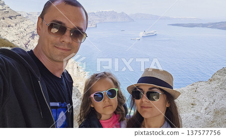 Family selfie at the iconic Heart of Santorini viewpoint overlooking the Aegean Sea caldera, cliffside villages and anchored cruise ship on a sunny Mediterranean day Family selfie at the iconic Heart of Santorini viewpoint overlooking the Aegean Sea caldera, cliffside villages and anchored cruise ship on a sunny Mediterranean day 137577756