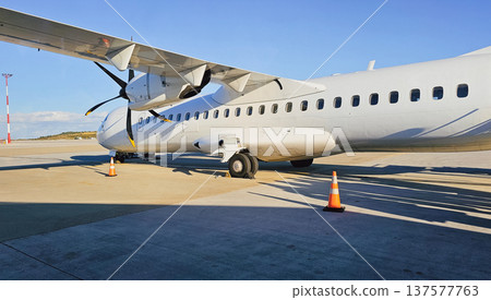 Mid-section of white passenger turboprop aircraft on sunlit tarmac with six-blade propeller, wing structure, landing gear, and runway under clear blue sky 137577763