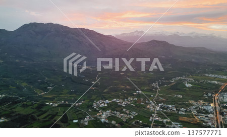 Aerial panoramic view of Crete foothills at sunset with snow-capped peaks, olive groves, valley with lake, scattered villages, winding road, and layered clouds Aerial panoramic view of Crete foothills at sunset with snow-capped peaks, olive groves, valley with lake, scattered villages, winding road, and layered clouds 137577771