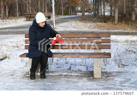 Woman sitting on bench and pouring drink from thermos while enjoying winter day Woman sitting on bench and pouring drink from thermos while enjoying winter day 137579607
