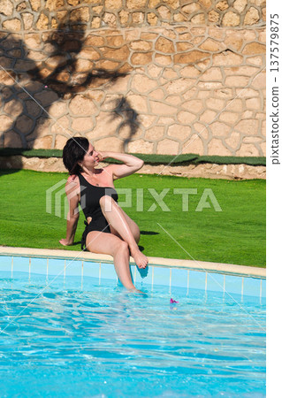 A woman in a bathing suit sits on the grass near a swimming pool with blue tiles 137579875