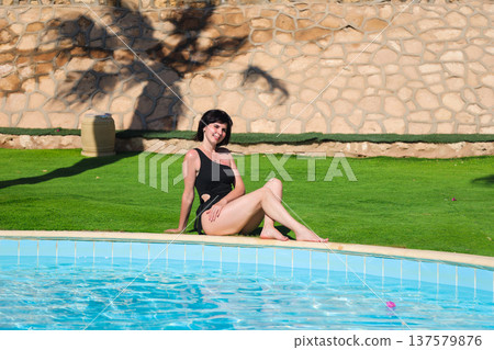 A woman in a bathing suit sits on the grass near a swimming pool with blue tiles 137579876