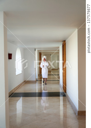 A woman in a white robe walks down a long hotel corridor 137579877