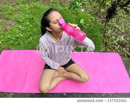 Young Filipino woman drinking water from pink bottle after yoga workout, Asian lady resting on mat in tropical garden, fitness lifestyle and wellness Young Filipino woman drinking water from pink bottle after yoga workout, Asian lady resting on mat in tropical garden, fitness lifestyle and wellness 137581081