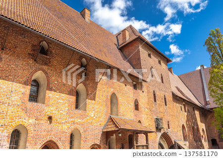 The Malbork Castle, a brick Gothic castle in northern Poland, Europe. Majestic medieval castle facade featuring red brickwork, arched windows, and tile roofing against a backdrop of blue skies. 137581170