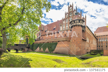 The Malbork Castle, a brick Gothic castle in northern Poland, Europe. Majestic brick castle with ornate architecture, surrounded by lush greenery and a blue sky with scattered clouds. The Malbork Castle, a brick Gothic castle in northern Poland, Europe. Majestic brick castle with ornate architecture, surrounded by lush greenery and a blue sky with scattered clouds. 137581175