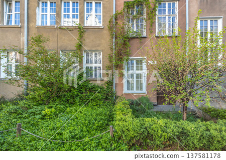 Gdansk, a city on the Baltic coast of northern Poland, Europe. Facade of building with climbing plants, green bushes, and a small tree. The windows have white frames, some show reflections. Gdansk, a city on the Baltic coast of northern Poland, Europe. Facade of building with climbing plants, green bushes, and a small tree. The windows have white frames, some show reflections. 137581178