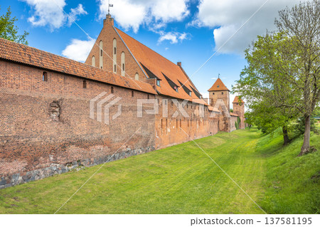The Malbork Castle, a brick Gothic castle in northern Poland, Europe. Majestic castle with brick walls, red roof, and vibrant green grass under a beautiful sky with clouds. Historic architecture. 137581195