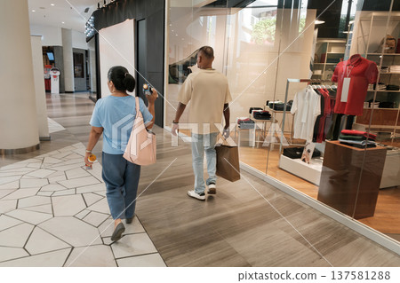 Two people walk past a store in a shopping mall carrying items during the day in a busy retail space Two people walk past a store in a shopping mall carrying items during the day in a busy retail space 137581288