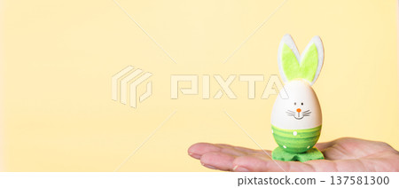 Decorative Easter egg with bunny ears held in hand against yellow background 137581300
